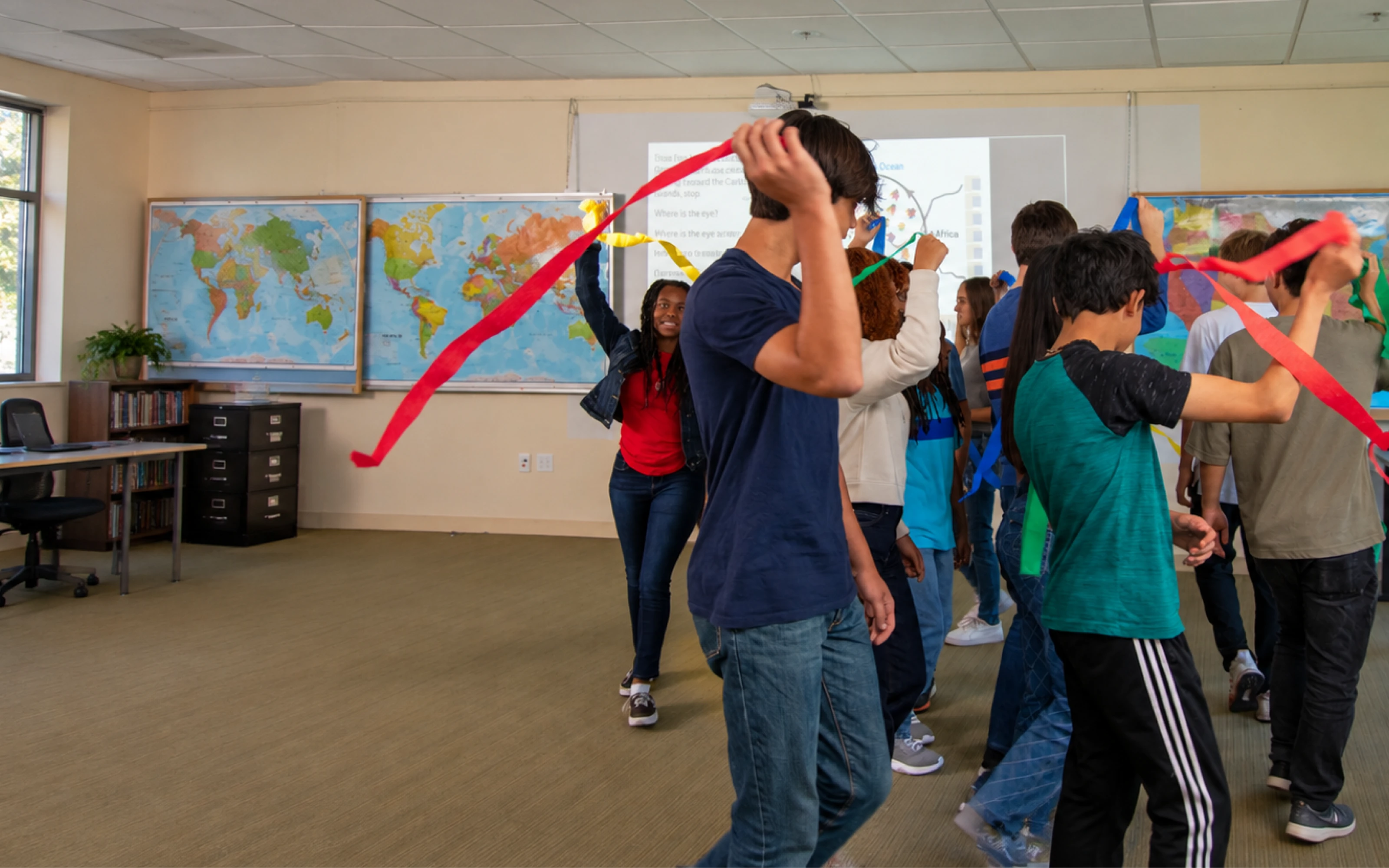 Students in a classroom dance with colorful streamers; world maps hang on the classroom walls in the background.