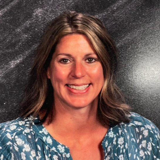 Smiling woman with shoulder-length brown hair in a blue floral top, posed in front of a gray background.