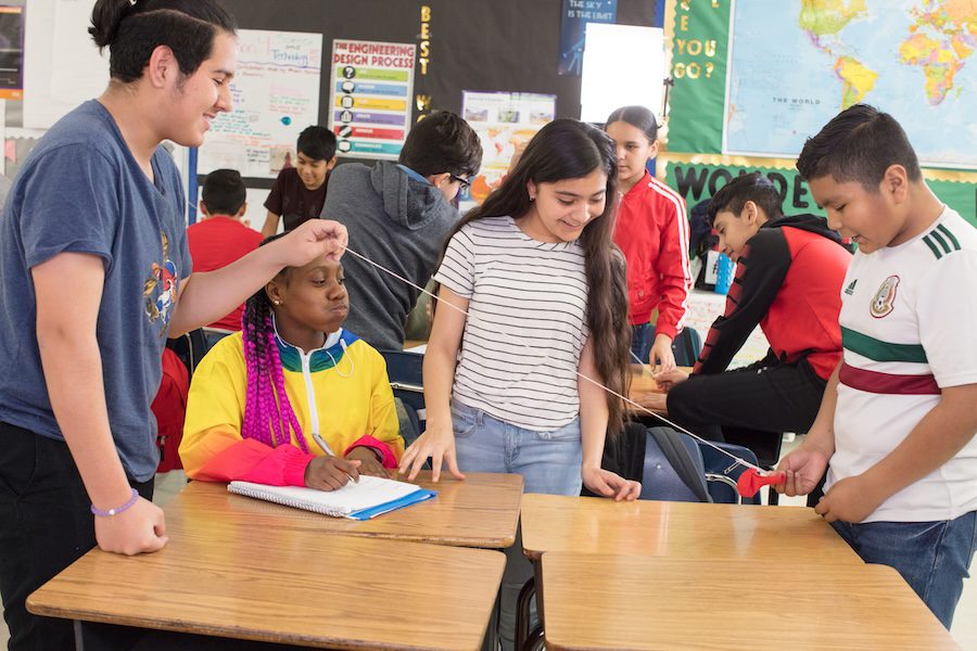 Four students use string in a classroom activity while other students work together in the background.
