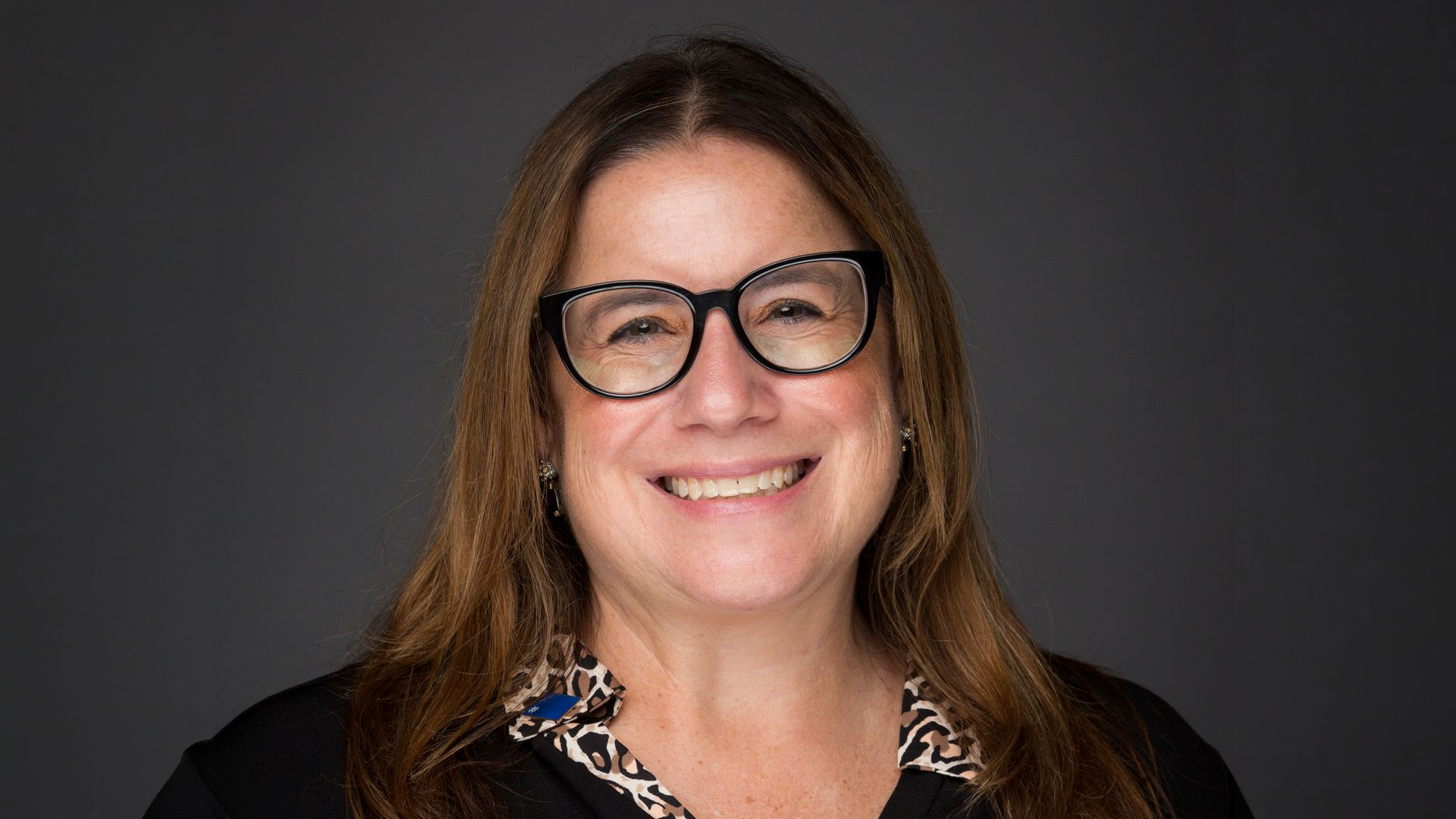 Woman with long brown hair, black glasses, and a patterned collar smiling against a dark background.