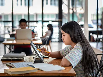 A student uses a stylus on a tablet at a table with books, with other students studying in the background.