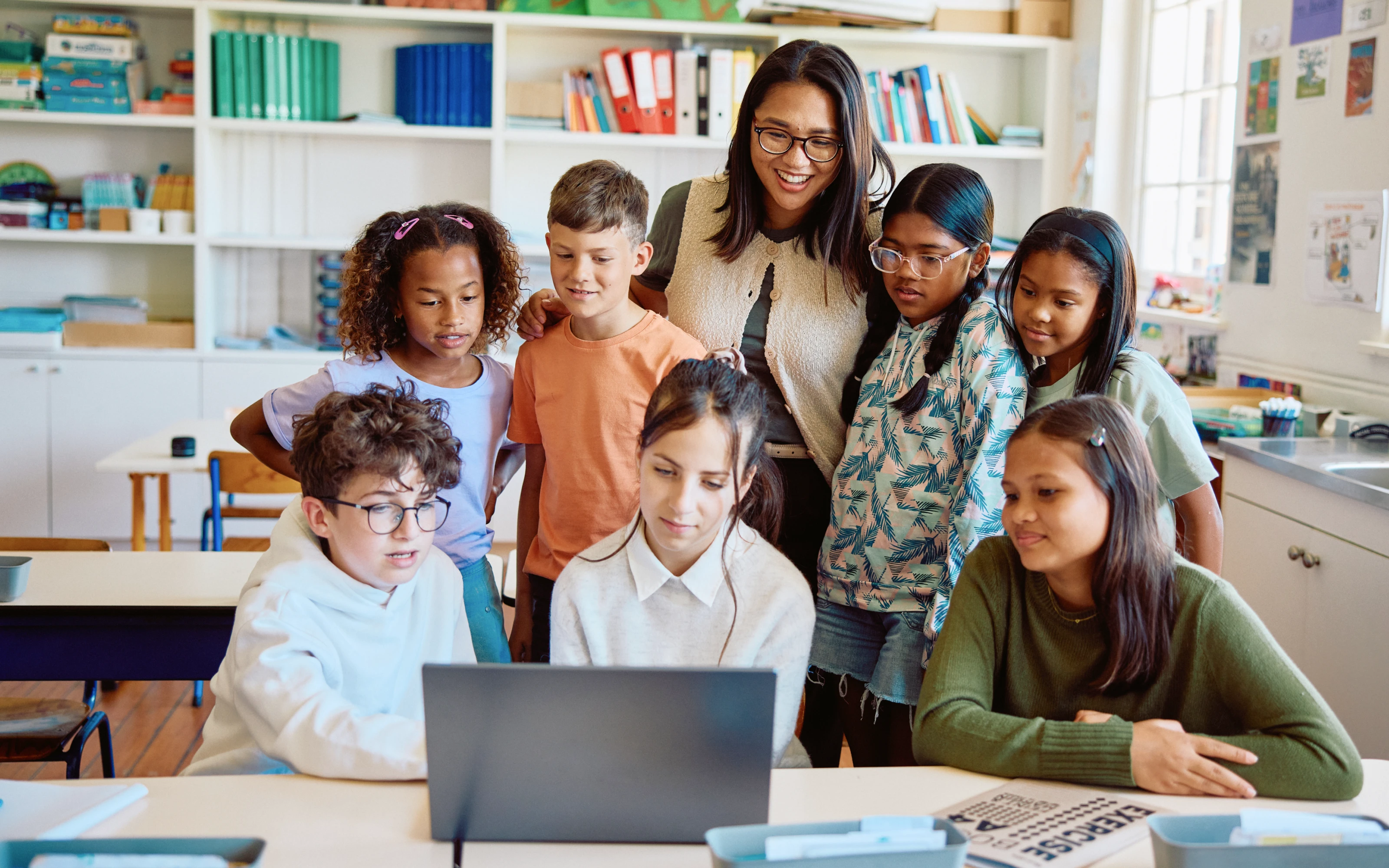 A teacher and seven students gather around a laptop, smiling and looking at the screen in a classroom.