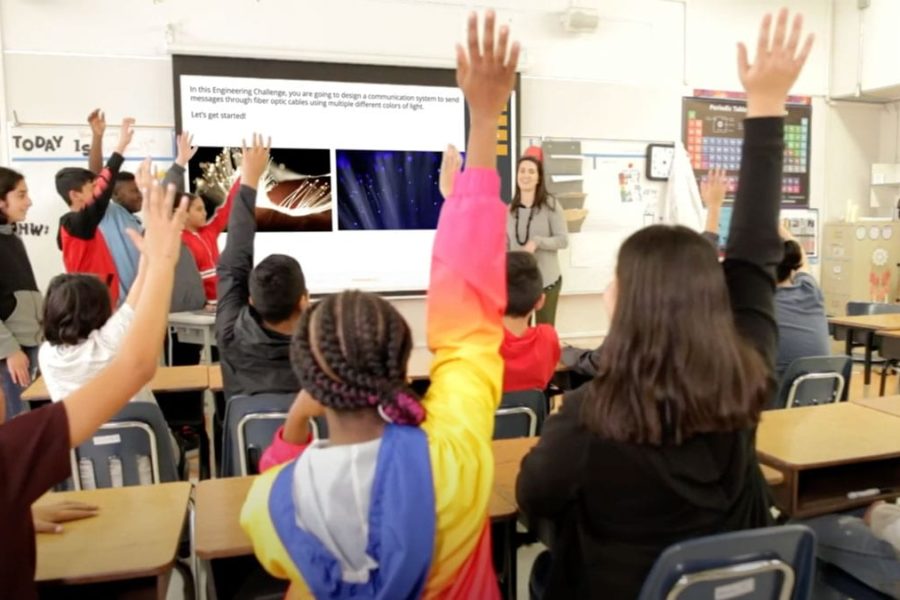 Students in a classroom raise their hands while a teacher stands at the front near a screen with images and text displayed.