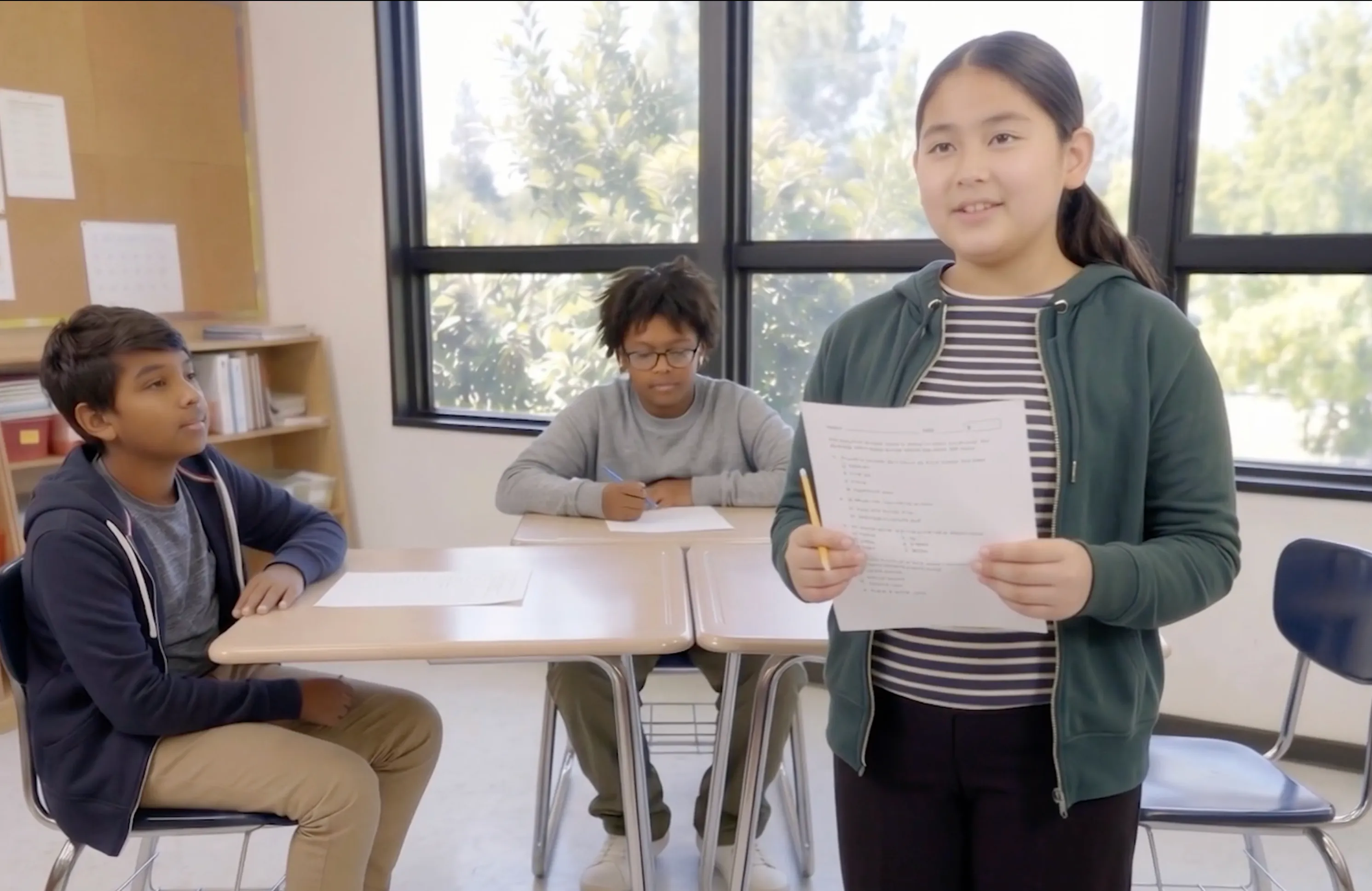A student stands holding a paper, presenting to two classmates seated at desks in a bright classroom.