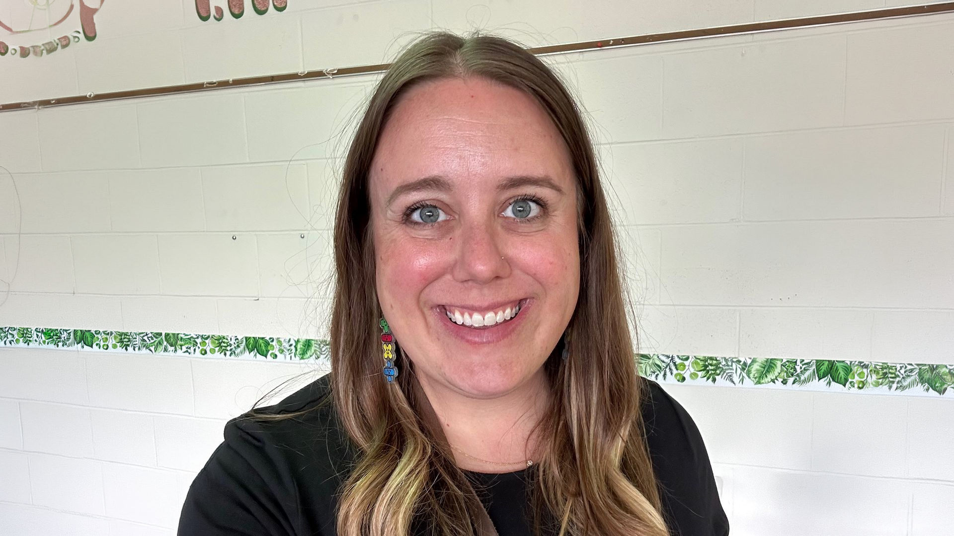 Woman with long brown hair smiling, wearing colorful earrings and a black shirt, standing in front of a white wall.