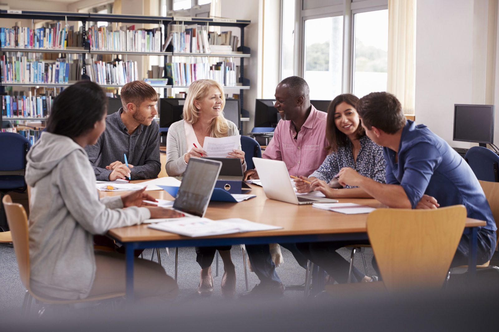 Six people sit around a table in a library, smiling and talking, with laptops and papers in front of them.