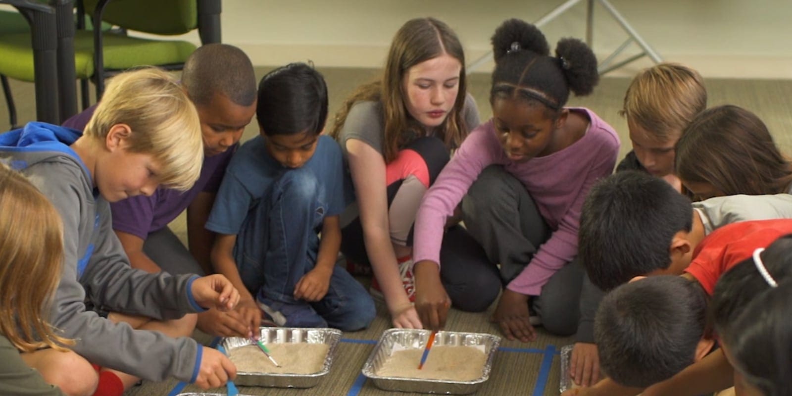 A group of children sit in a circle, using brushes to dig in trays filled with sand placed on the floor.