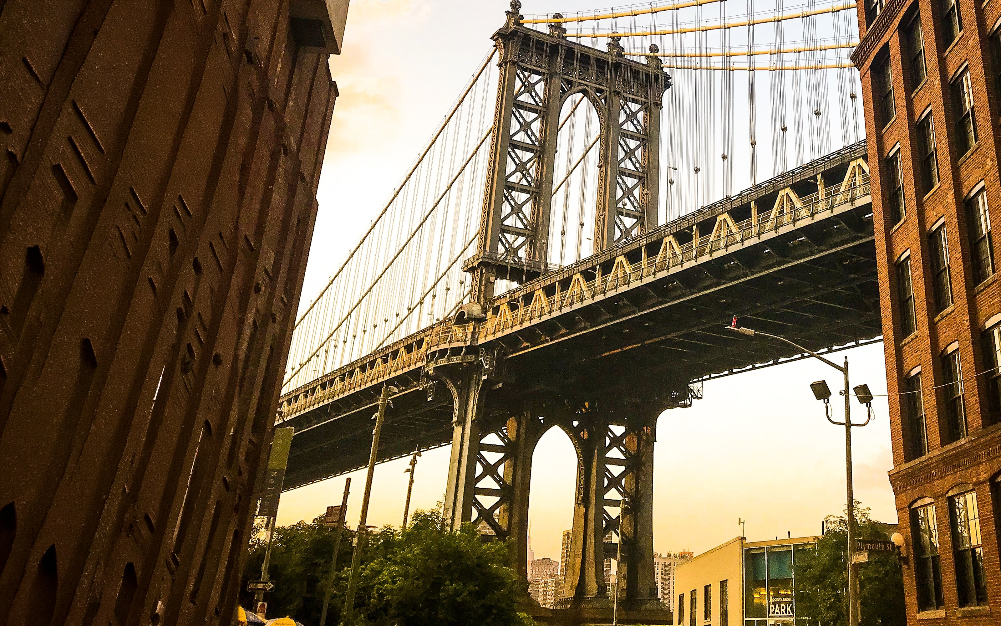Manhattan Bridge seen between two brick buildings at sunset, with trees and streetlights below.
