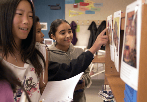 Three students look at and point to images on a shelf while holding notebooks in a classroom setting.