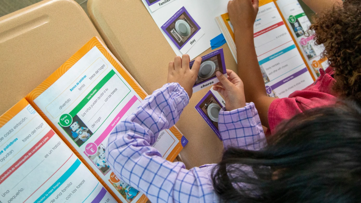 Two children sit at desks, working with colorful educational worksheets and cards featuring pictures and words.