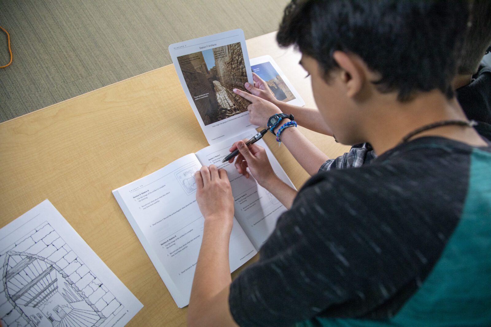 Two students work together, referencing a booklet and writing in a workbook on a light wood table.