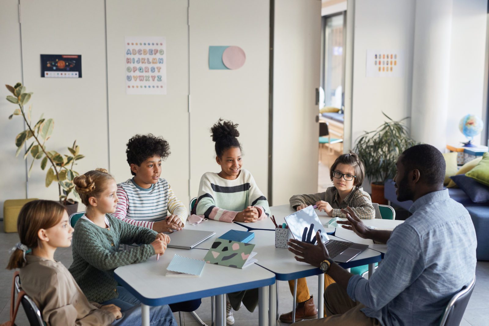A teacher sits at a table with five students in a classroom, discussing a laptop screen.