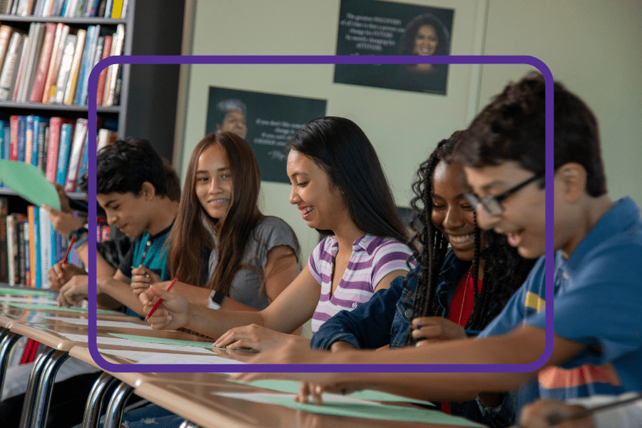 A group of diverse students sitting at desks in a classroom, smiling and working on papers.