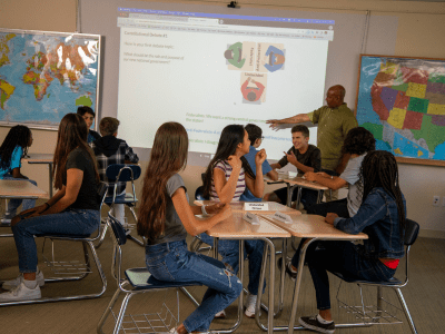 Students sit at desks in a classroom while a teacher points to a projection on the whiteboard.