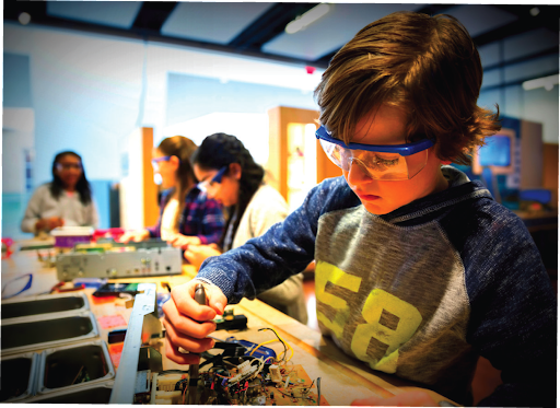 A young student wearing safety goggles uses a screwdriver to work on a disassembled electronic device in a classroom.