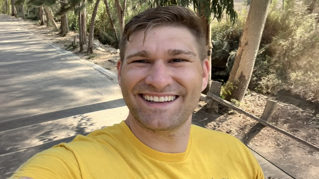 Smiling man in a yellow shirt takes a selfie on a sunny path lined with trees and greenery.