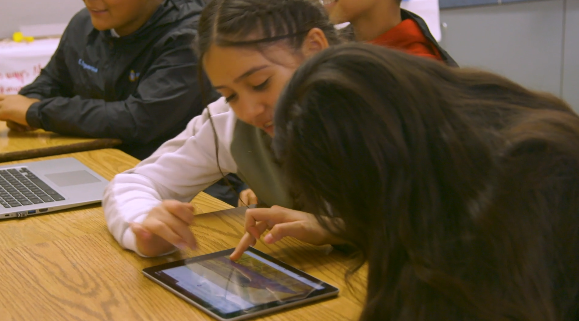 Two students use a tablet together at a wooden desk, smiling and engaged, with a laptop nearby.