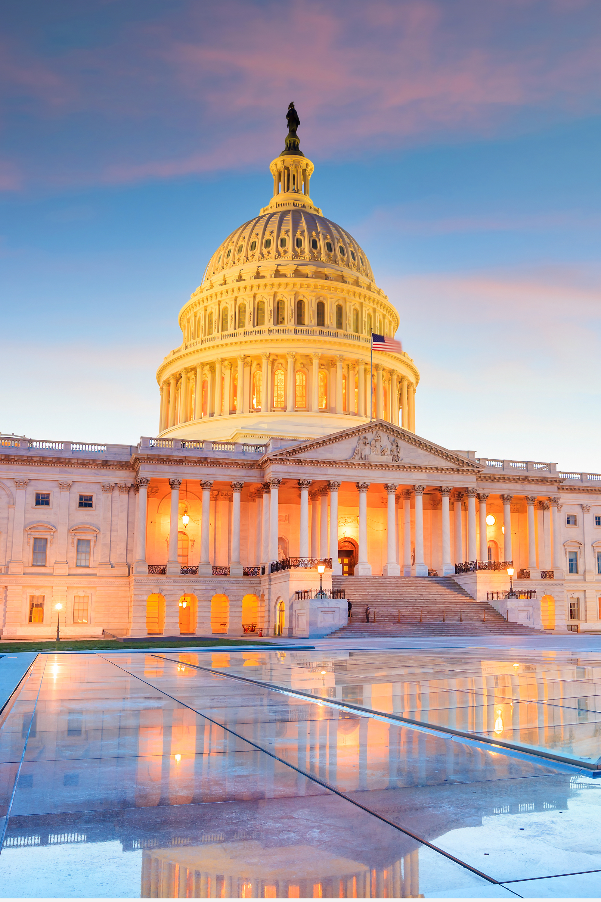The U.S. Capitol building at sunset with lights glowing and its reflection in a pool in front.