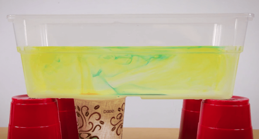 Clear container with yellow and green liquid, resting on red plastic cups and a paper cup underneath.