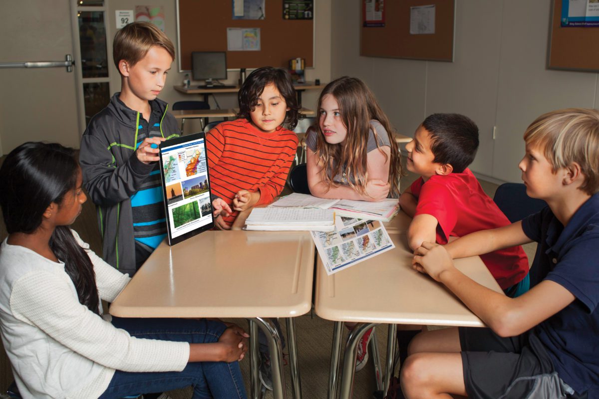Six children sit around desks while one child shows images on a tablet to the group in a classroom.
