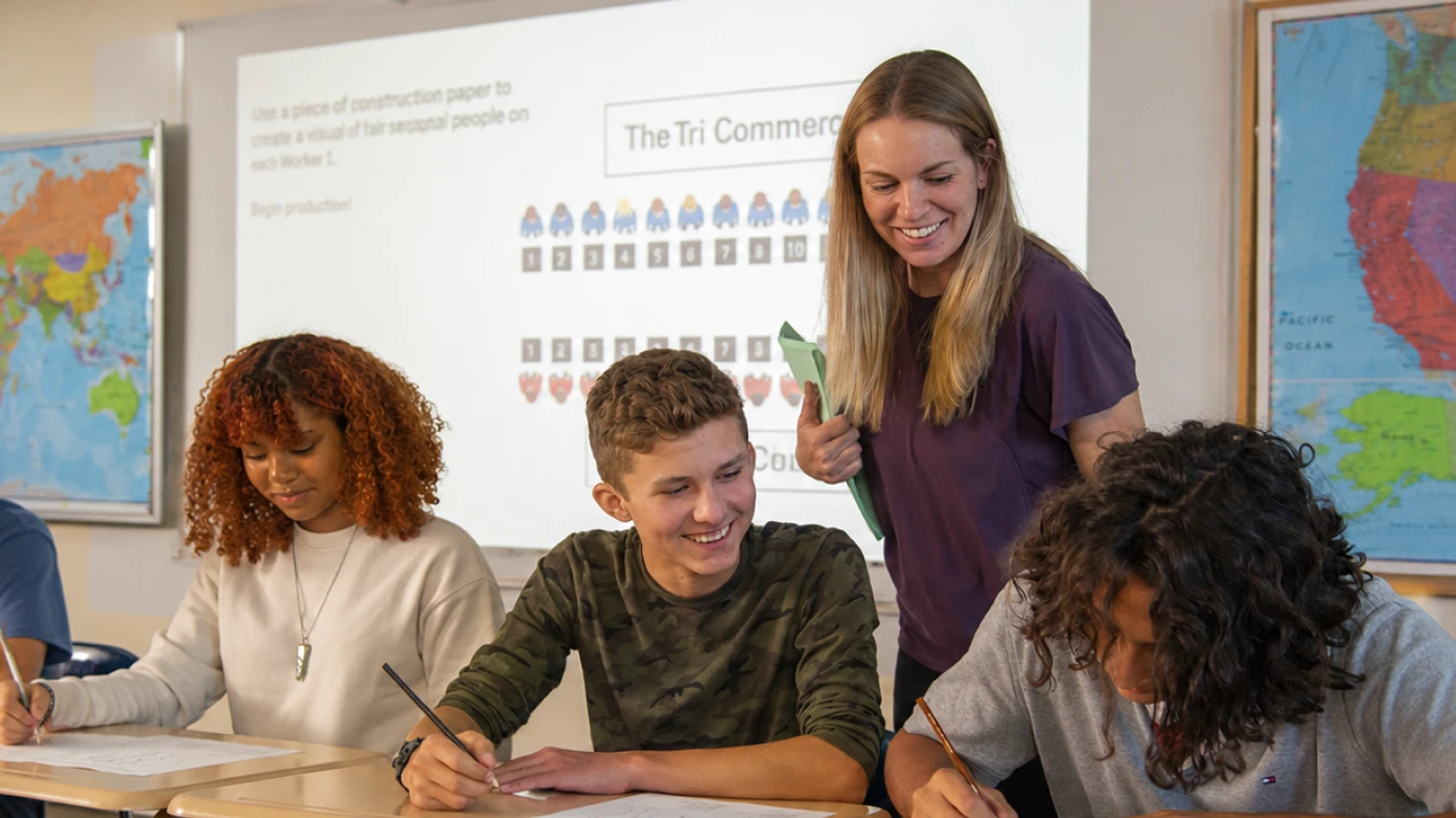 A teacher stands beside three students working at their desks in a classroom with world maps on the walls.