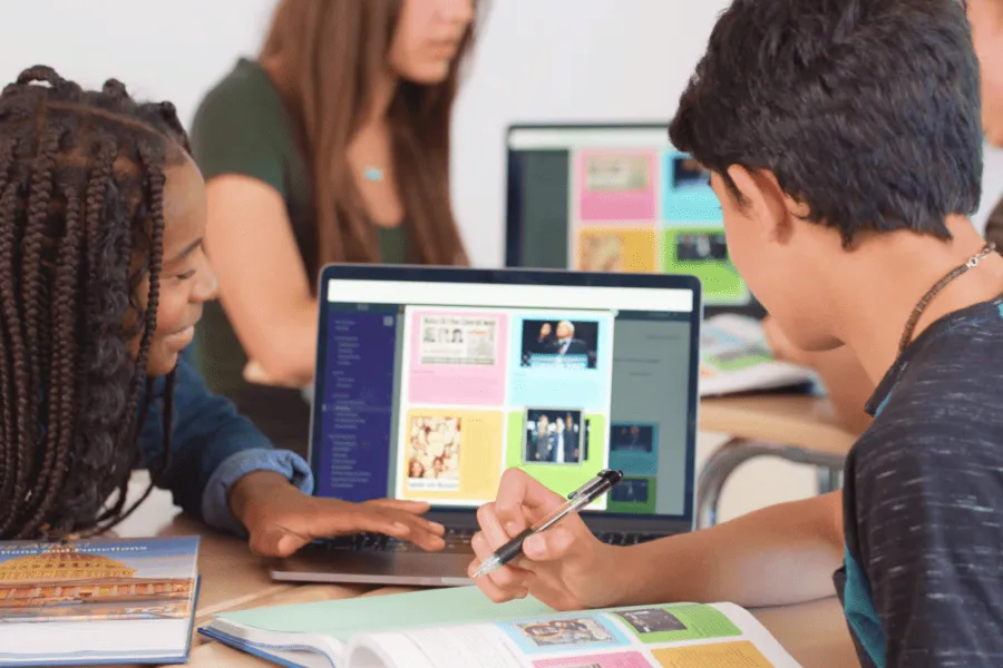 Two students collaborate at a desk, looking at a colorful website on a laptop in a classroom setting.