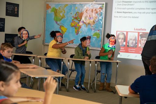 Five students wearing face masks act out a scene in a classroom with a world map and a projected slide.