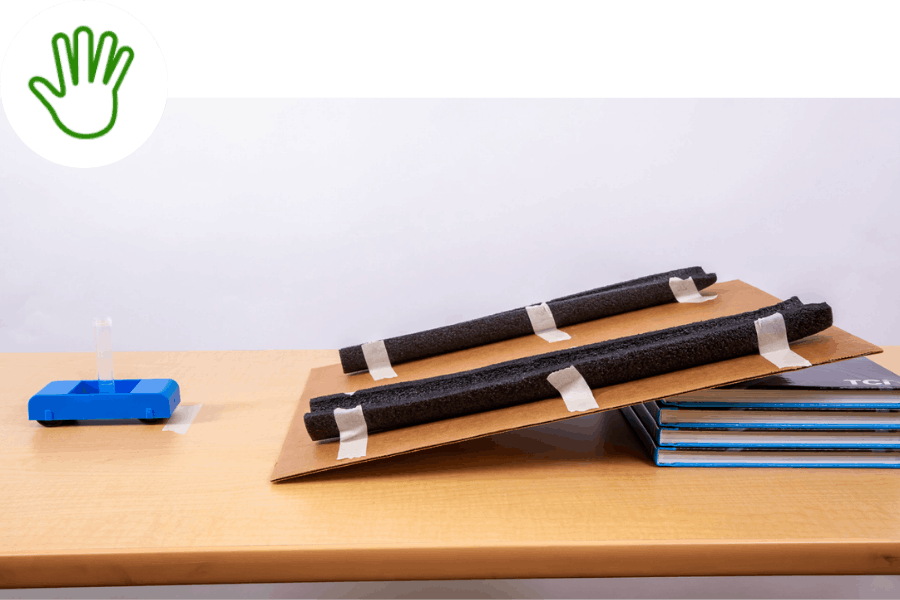 Inclined cardboard ramp with foam rails taped on, next to stacked books and a blue dispenser on a wooden table.