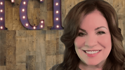 A smiling woman stands in front of a wooden wall with large, illuminated purple letters.