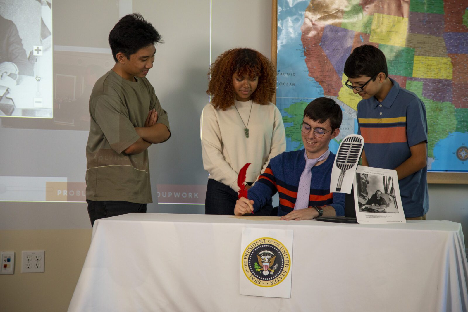 Four students gather around a table with a presidential seal; one signs a document with a red feather pen.