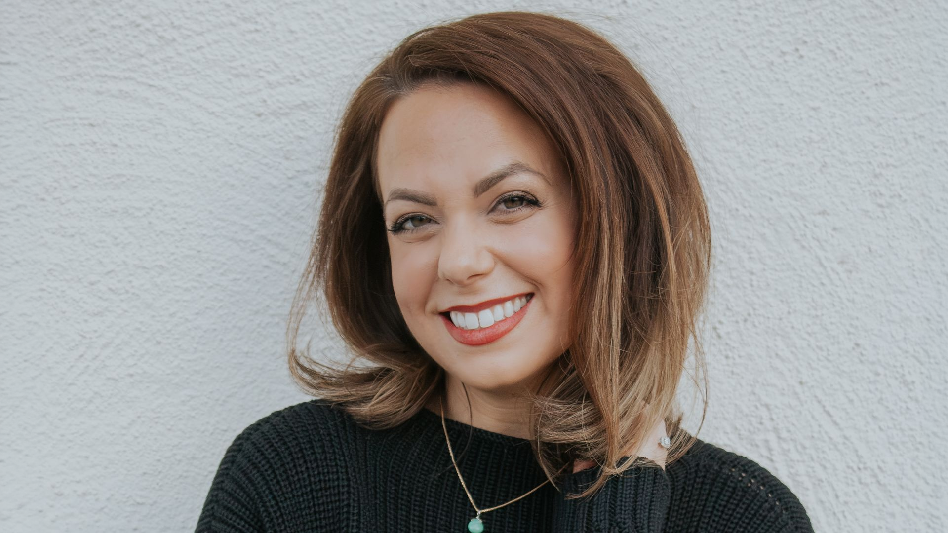 Woman with brown hair wearing a black sweater and necklace, smiling in front of a textured white wall.