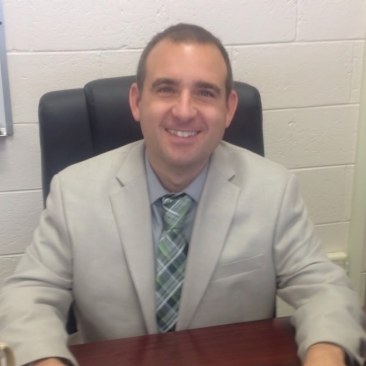 Man in a light gray suit and patterned tie smiles while sitting at a desk in an office.