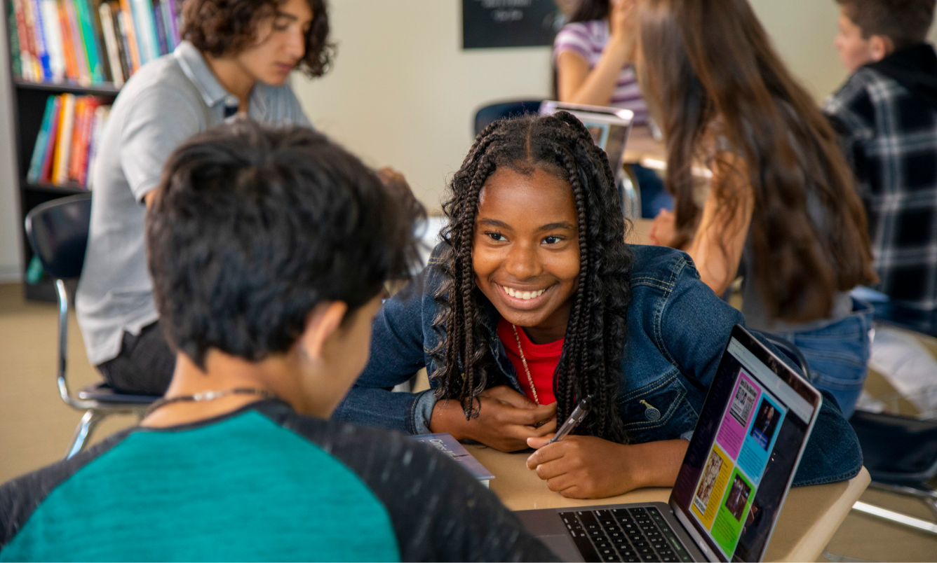 A girl smiles while talking to a classmate. They sit at a desk with a laptop. Other students are in the background.