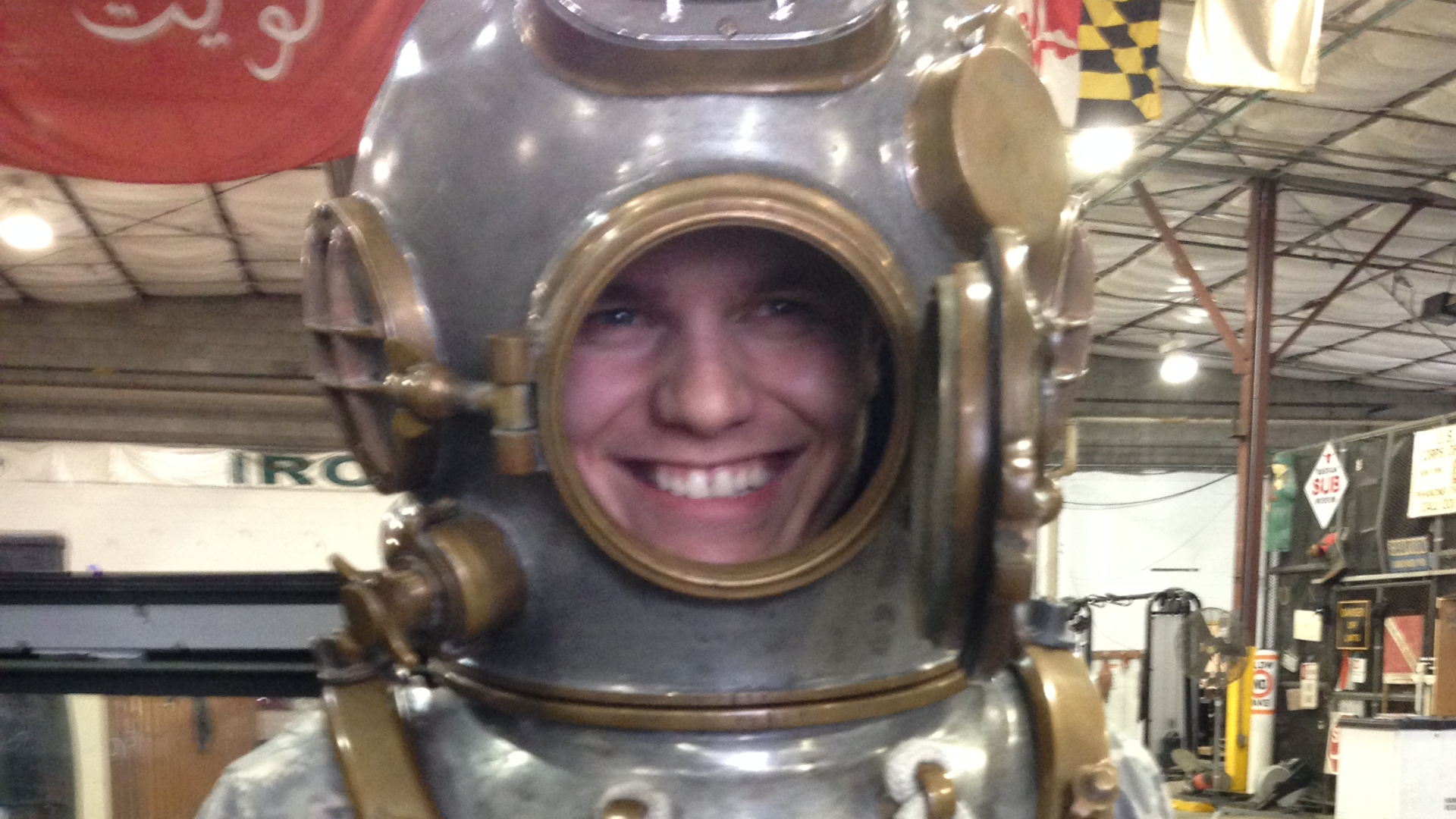 A smiling person wearing a vintage metal diving helmet in an indoor space with flags and industrial decor.