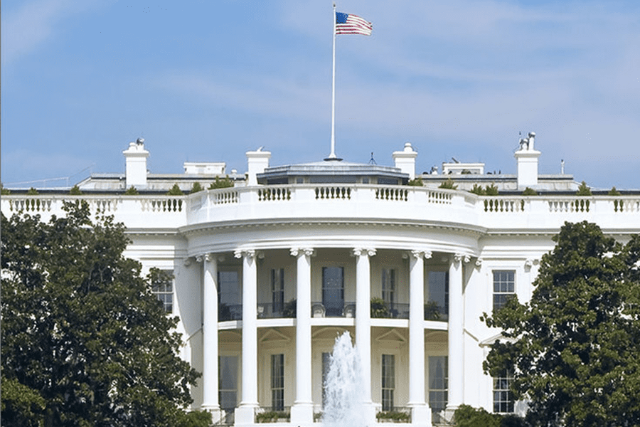 The White House with an American flag flying above and a fountain in front, surrounded by green trees.