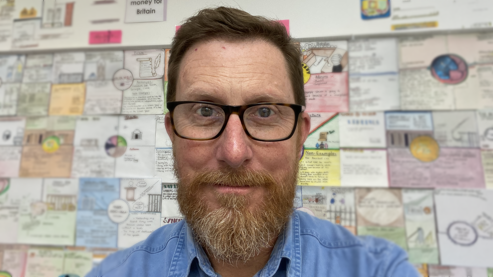 A man with glasses and a beard smiles in front of a colorful wall covered in drawings and notes.