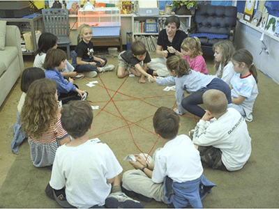 Twelve children and an adult sit in a circle on the floor, connecting yarn strands between them.