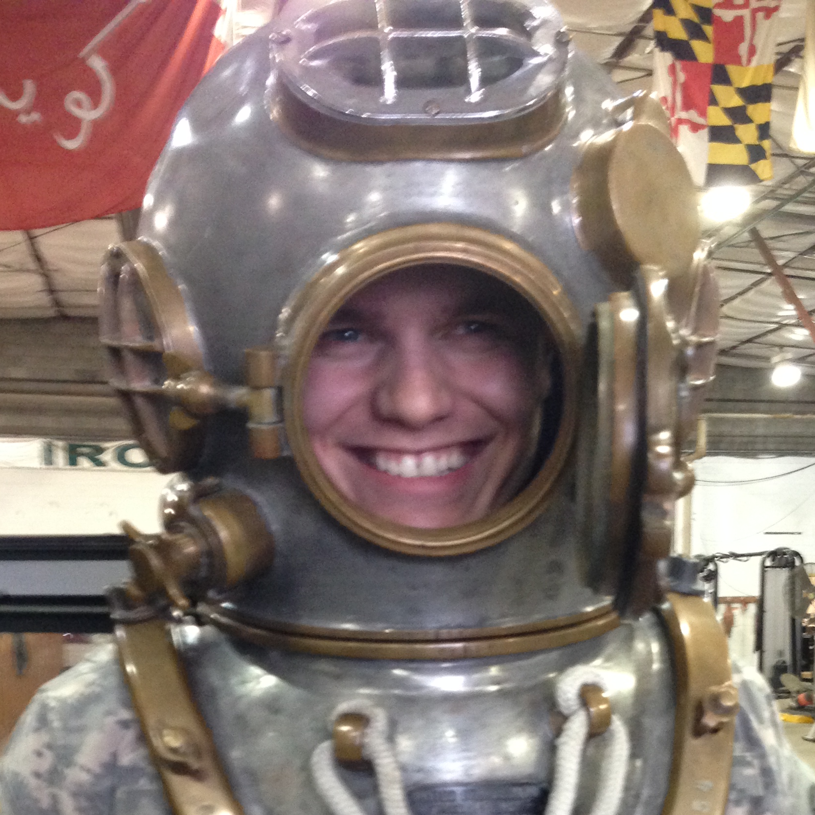 Smiling person inside a vintage metal deep-sea diving helmet at an indoor event or exhibition.