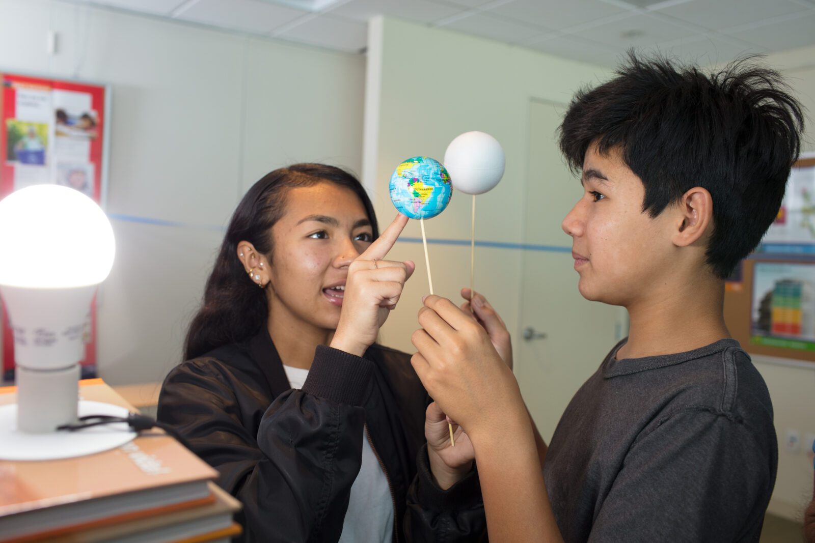 Two students holding models of the Earth and Moon to demonstrate an eclipse using a lamp.