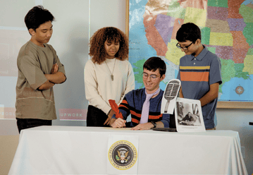 Four students reenact a presidential signing at a table with a mock seal and historical photo.