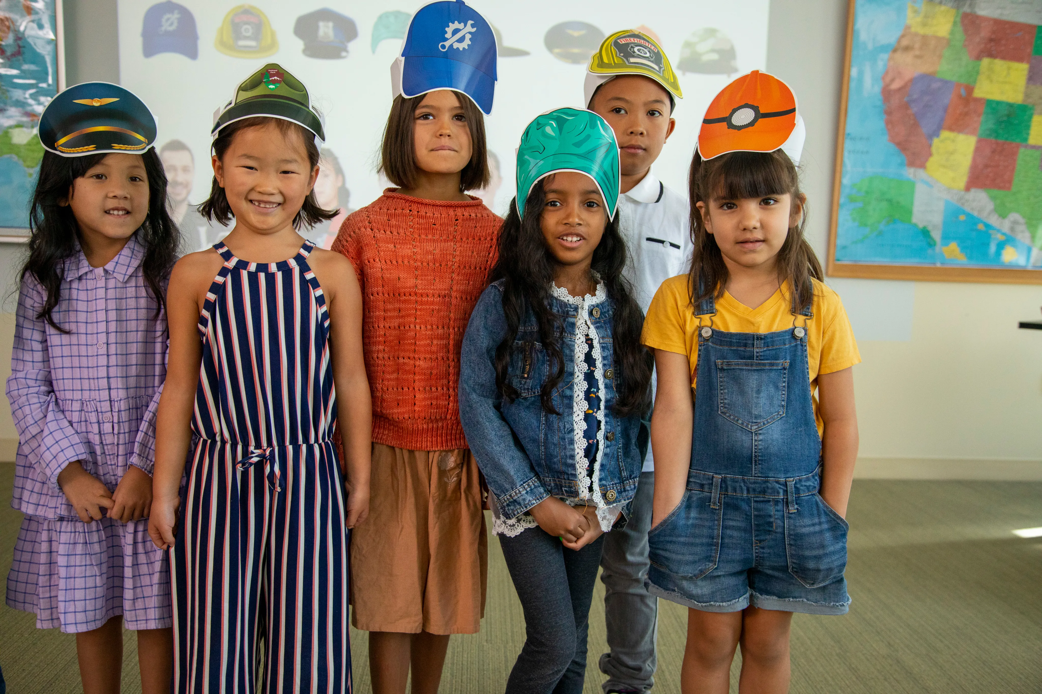 Six children wearing costume hats and casual clothes stand together, smiling in a classroom.