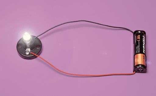 A glowing LED connected to a battery with red and black wires, forming a simple electrical circuit on a purple background.