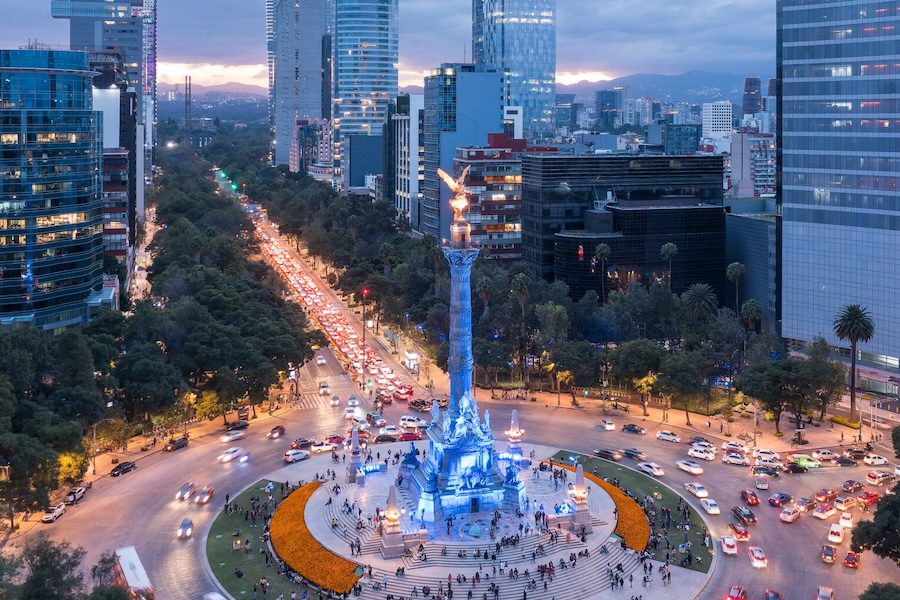 Aerial view of the Angel of Independence monument surrounded by city buildings and traffic in Mexico City at dusk.