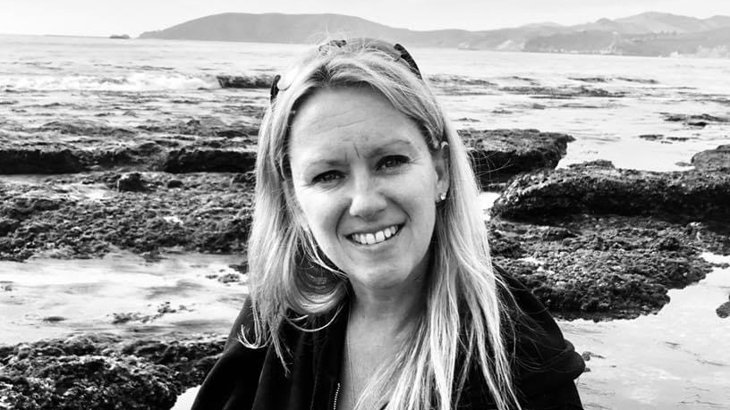Smiling woman with long hair sits by rocky shore, with ocean and hills in the background. Black and white photo.