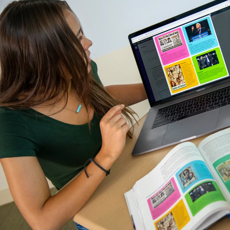 A student looks at a colorful history website on a laptop while studying from an open textbook at a desk.