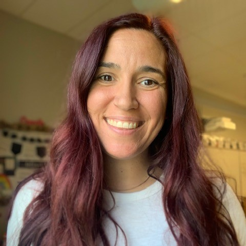 Smiling person with long, wavy dark red hair wearing a white top, indoors with a blurred background.