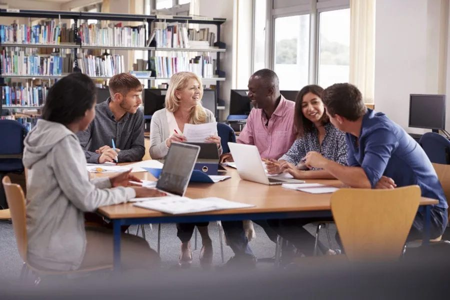 Six people sit around a table with laptops and papers, talking and smiling in a library setting.