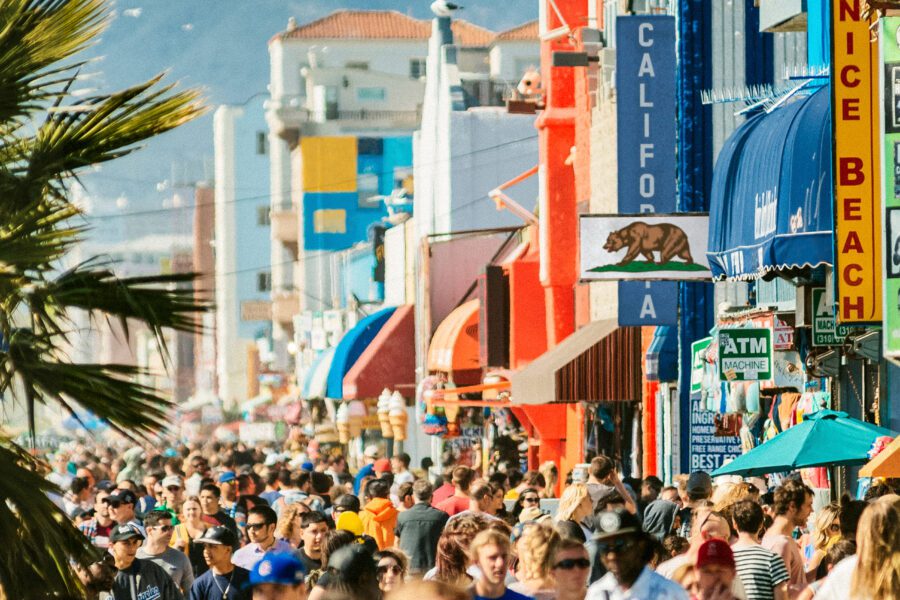 Crowded boardwalk lined with colorful shops and palm trees at Venice Beach, California, on a sunny day.
