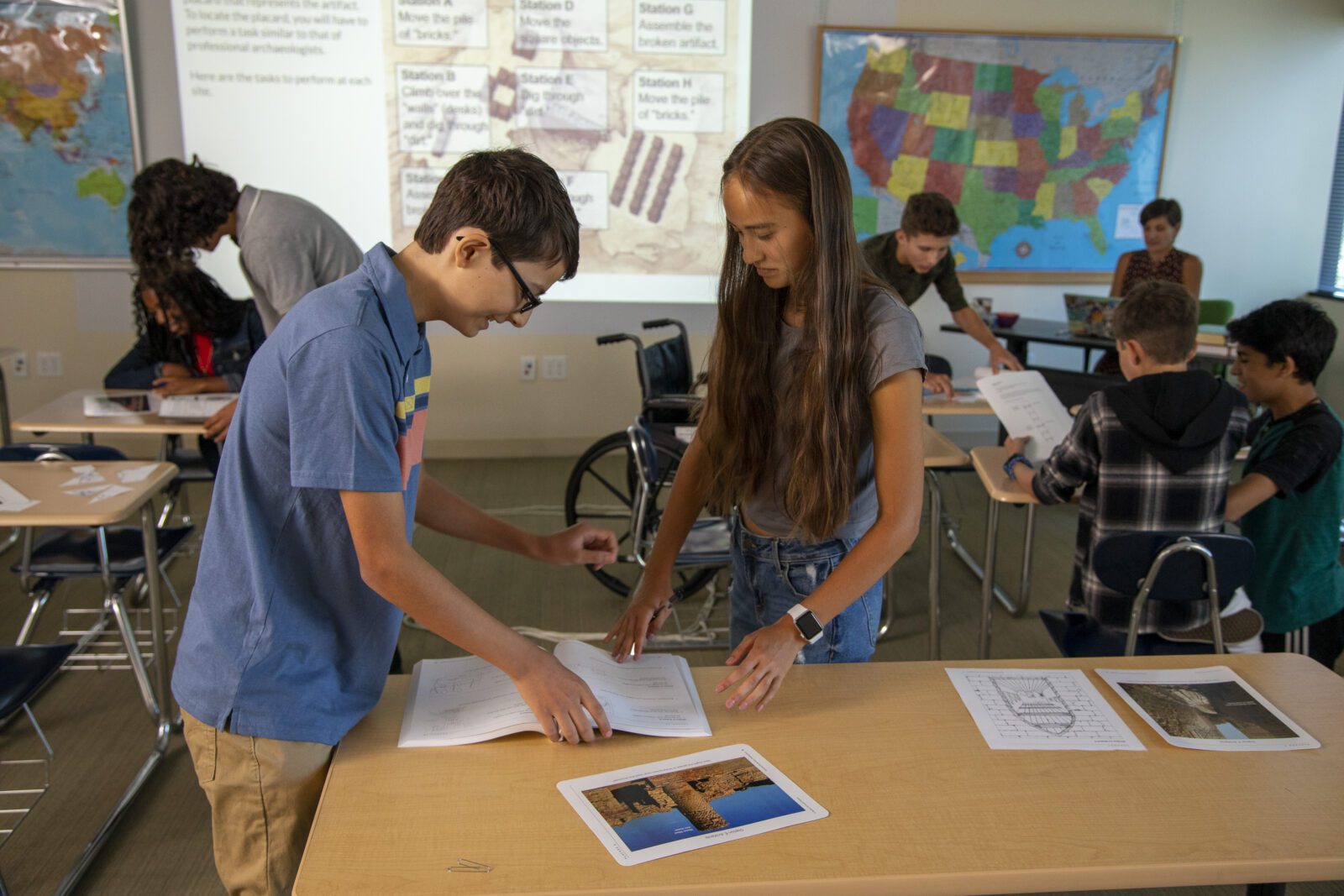 Two students work together at a table looking at papers, with other students and a map visible in a classroom.