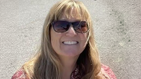 Woman with long blonde hair and sunglasses smiling outdoors on a sunny day.